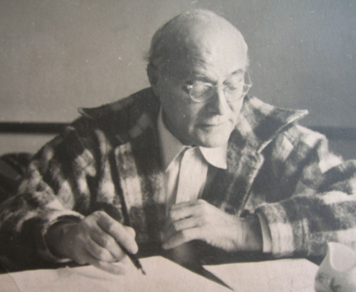 Man wearing glasses and plaid jacket sitting at a desk, writing.