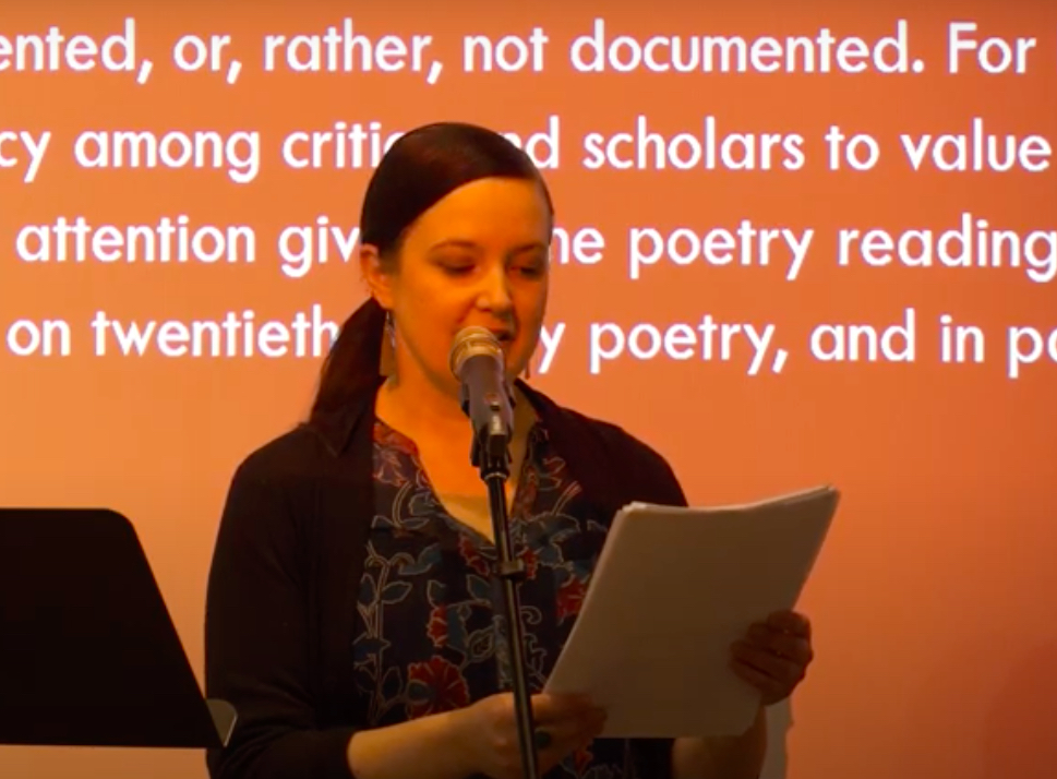 Katherine McLeod speaking in front of microphone with presentation in background