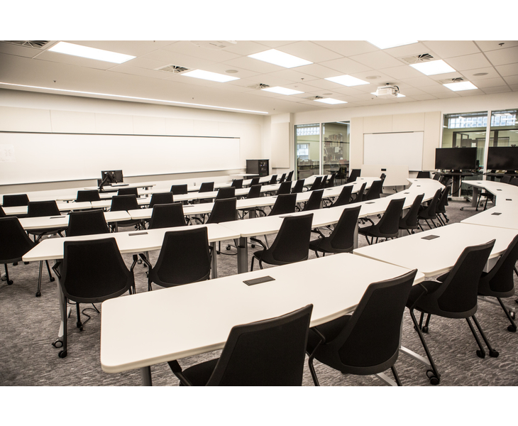 Large white-walled room with white tables and and chairs arranged in arcs facing the front of the classroom