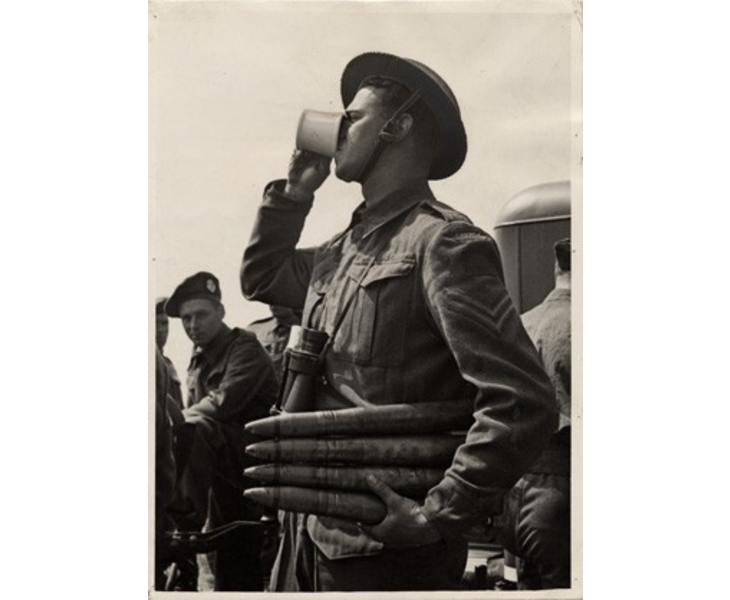 Soldier seen in profile drinking a cup of coffee, holding missiles in his left arm.